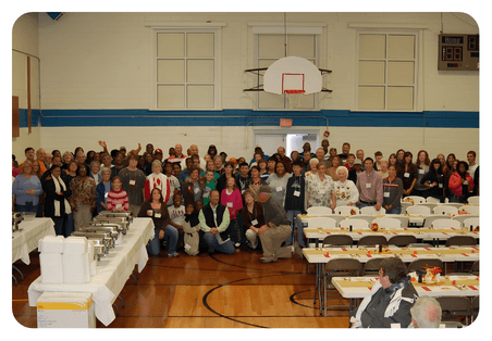 Large group of people posing in a gymnasium set up for a community dinner.