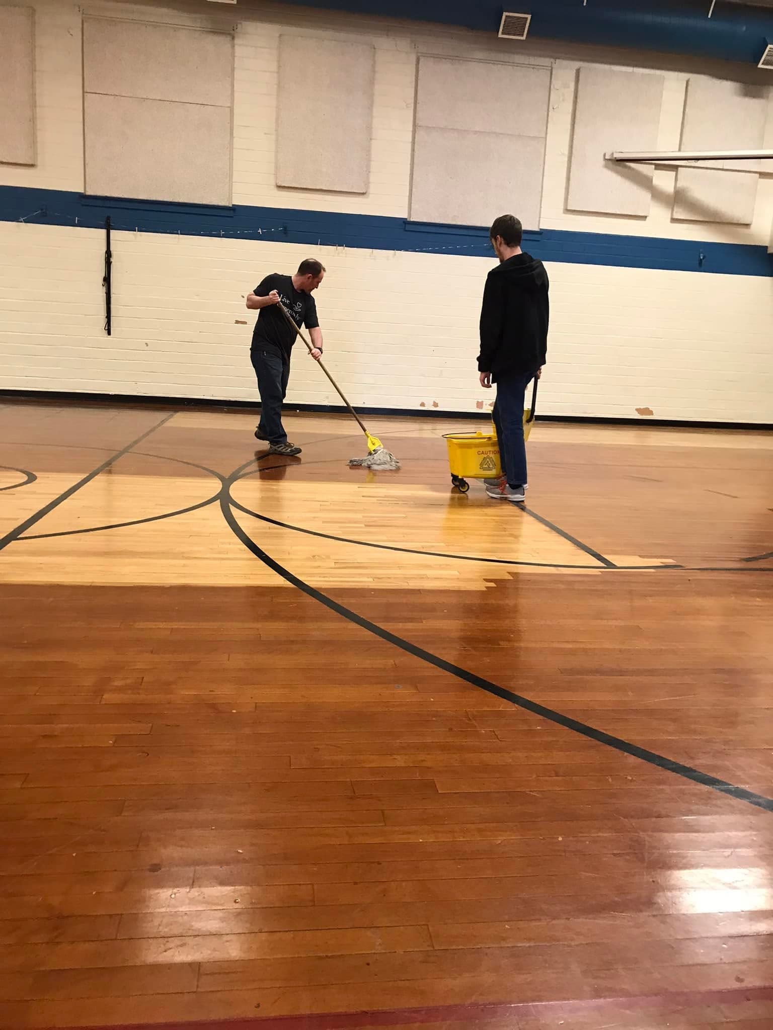 Two men cleaning a polished wooden basketball court floor with a mop and yellow bucket.