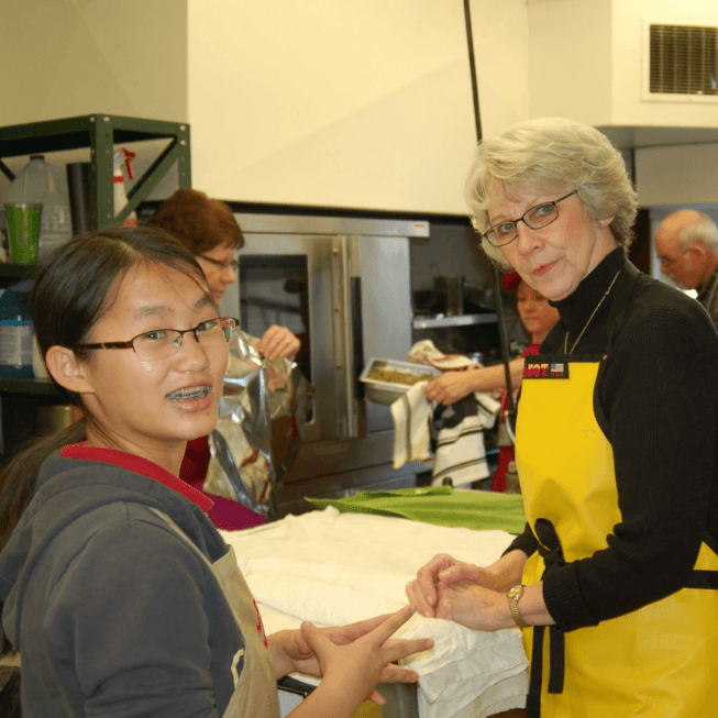 Young girl and woman in yellow apron smile while preparing food in a commercial kitchen.
