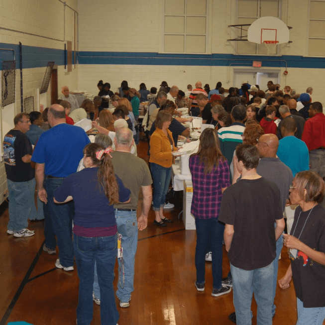 Diverse group of people lining up for food at tables inside a school gymnasium.