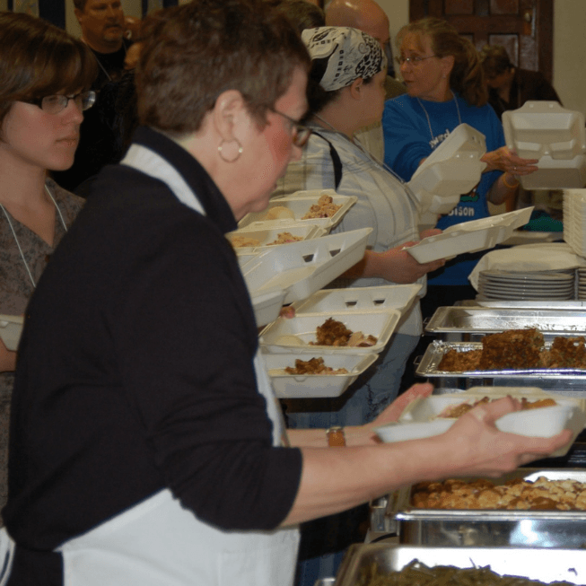 People filling white styrofoam containers with food from a buffet line of large metal trays.
